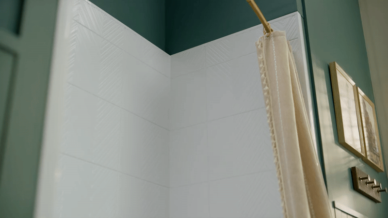 Close-up of Bath Fitter seamless white acrylic wall panels in a shower corner with teal painted walls, brass curtain rod, and cream shower curtain