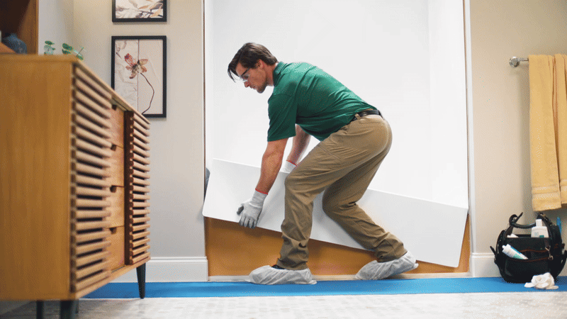Bath Fitter installer in green uniform and protective gear fitting a white acrylic bathtub liner in a residential bathroom with wood cabinet and protective floor covering